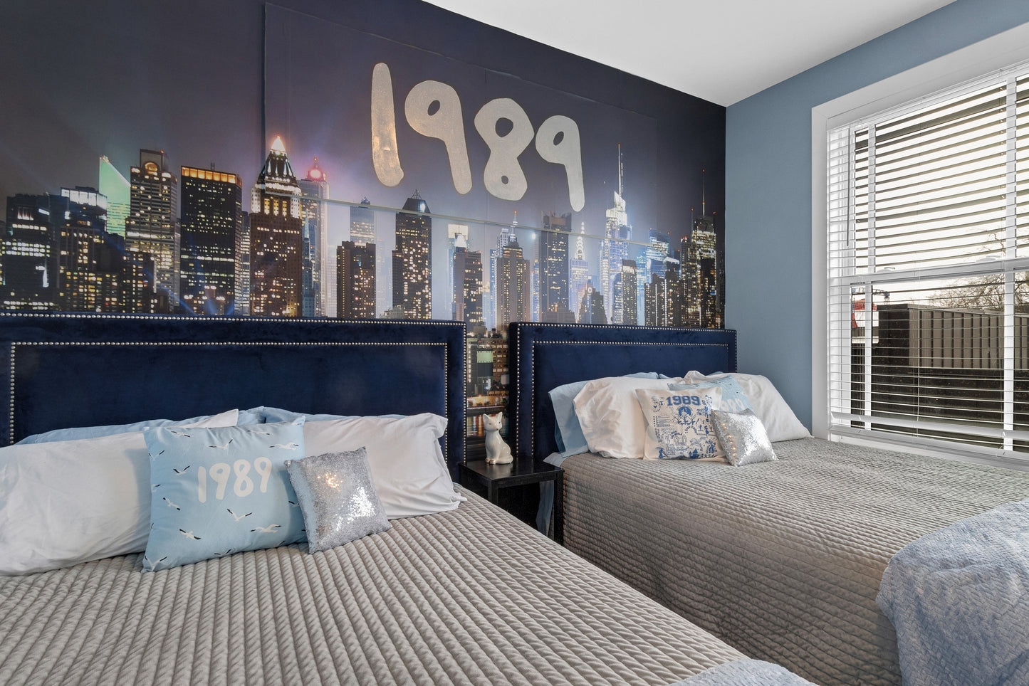 Two beds in a bedroom with a cityscape mural on the wall featuring the year 1989.